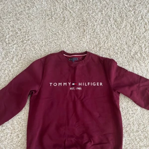 Vinröd sweatshirt från Tommy Hilfiger - Säljer en vinröd sweatshirt från Tommy Hilfiger med klassisk logga och text framtill. Tröjan har rund halsringning, långa ärmar och är tillverkad i mjuk bomull. Perfekt för en avslappnad och stilren look.