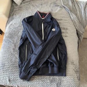 Mörkblå vindjacka från Moncler - Snygg mörkblå vindjacka från Moncler med vita detaljer och dragkedjor. Jackan har Moncler-logga på ärmen, ribbade muddar och hög krage med rödvit rand. Tillverkad i 100% polyamid, perfekt för vår och höst. Klassisk och sportig look.