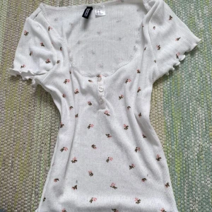 Vit blommig topp från H&M Divided XS - Supersöt vit topp från H&M Divided i storlek XS med små rosa blommor, korta ärmar och knappar framtill. Toppen har lätt ribbad struktur och en feminin, något figurnära passform. Perfekt för en romantisk look!
