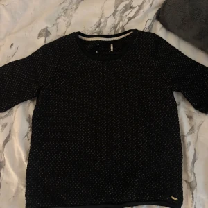 Svart mönstrad tröja Holly & White - Säljer en svart tröja från Holly & White, Lindex  med diskret rutigt mönster i guldtråd. Tröjan har rund halsringning och trekvartsärm. Perfekt för dig som gillar stilrena detaljer och en lite lyxigare känsla. Storlek xs/s