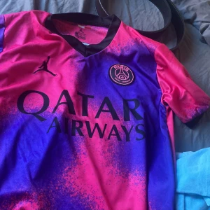 PSG Mbappé fotbollströja Jordan - Paris Saint-Germain fotbollströja med Mbappé 7 på ryggen. Tröjan har en fet mix av rosa och blå färg med svart logga och detaljer, samt Qatar Airways tryck på bröstet. Officiell Ligue 1-patch på ärmen. Tillverkad i lätt och ventilerande polyester.