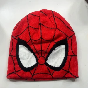Röd Spiderman-mössa med nätmönster - Cool röd mössa med Spiderman-design, stora vita ögon och svarta spindelnätslinjer. Perfekt för dig som gillar Marvel och vill sticka ut i vinter. Tillverkad i mjukt stickat material som håller dig varm.