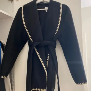 Svart kappa med bälte och mönster - Svart lång kappa med bälte i midjan och dekorativa ljusa sicksack-detaljer längs krage, ärmslut och nederkant. Kappan har lång ärm och en oversized känsla, perfekt för att lyfta din outfit.