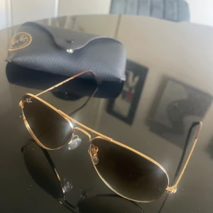 Ray-Ban aviator solglasögon! - Ray-Ban aviator solglasögon med tunna guldfärgade metallbågar och bruna tonade glas. Klassisk pilotform som ger en snygg och tidlös look. Levereras med svart Ray-Ban fodral med guldfärgad logga. Perfekt accessoar för soliga dagar.