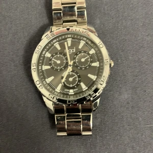 Silverfärgad klocka från Regal - Stilren klocka från Regal i silverfärgat rostfritt stål med länkarmband. Urtavlan är mörkgrå med tre mindre urtavlor och tydliga indexmarkeringar. Klockan har sportig design och metallisk finish, perfekt för dig som gillar klassisk stil med modern touch.