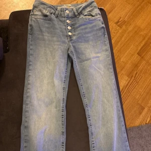 Midwaist jeans med knappar från Nelly - Snygga ljusblå midwaist jeans från Nelly med raka ben och fem dekorativa knappar framtill. Klassisk femficksmodell i mjukt denimtyg som ger en chill och avslappnad vibe. Perfekt för dig som gillar enkel och stilren look.