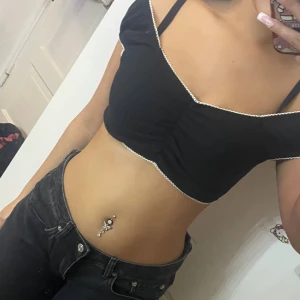 Svart croppad topp med spetskant - Säljer en svart croppad topp med v-ringning och vita spetsdetaljer längs kanterna. Toppen har en lätt rynkning framtill och är ärmlös med offshoulder-design. Perfekt för dig som gillar en söt och trendig stil.