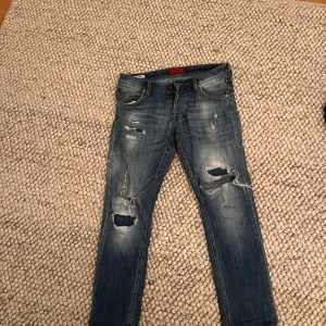 Blå slitna jeans från Jack & Jones - Säljer ett par blå jeans från Jack & Jones med snygga slitningar och hål på benen. Jeansen har klassisk femficksdesign, knappgylf och ljus tvätt med slitna detaljer både fram och bak. Perfekta för en avslappnad och trendig look. denna modell säljs inte längre 