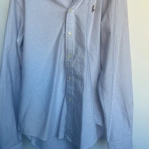 Ralph lauren skjorta - Säljer en klassisk vit och blå randig skjorta från Ralph Lauren med broderad logga på bröstet. Skjortan har lång ärm, knappar framtill och är i 100% bomull. Perfekt för dig som gillar stilrena och tidlösa plagg. Storlek S