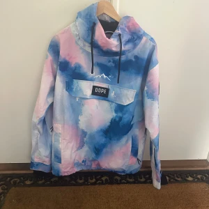 Dope Snow vindjacka i blått och rosa - Säljer en fet vindjacka från Dope Snow i storlek L med unikt blått, rosa och vitt mönster. Jackan har huva med snörning, stor ficka framtill med logopatch och coolt emblem på ärmen. Perfekt för dig som gillar street och färg. Säkra din stil nu!
