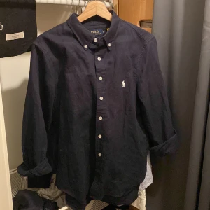 Mörkblå skjorta från Polo Ralph Lauren - Klassisk mörkblå skjorta från Polo Ralph Lauren i linne med button-down krage och vita knappar. Broderad logga på bröstet och relaxed passform. Perfekt för dig som gillar stilren och tidlös look.