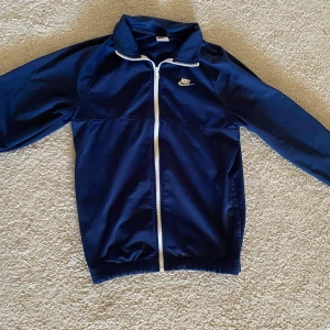 Blå vindjacka från Nike med dragkedja - Snygg marinblå vindjacka från Nike med vit dragkedja och klassisk logga på bröstet. Jackan har hög krage, långa ärmar och elastiska muddar. Perfekt för dig som gillar sportig stil och vill ha något lätt att slänga på sig.
