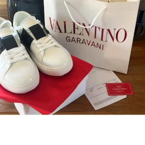 Valentino Garavani Open sneakers vit/svart - Använda exakt 1 gång då dom inte passar mig längre, köpta för 5800 (kvitto finns) Snygga Valentino Garavani Open sneakers i vitt skinn med bred svart läderdetalj över snörningen. Klassisk rund tå, platt sula med mönster och ikoniska nitar på hälen. Perfekt för dig som gillar exklusiv streetstyle och vill sticka ut med lyxiga detaljer.