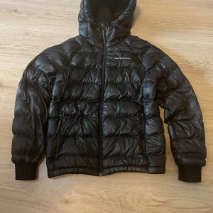 Svart dunjacka Peak Performance XS - Snygg svart dunjacka från Peak Performance i storlek XS. Jackan har huva, dragkedja framtill och två sidofickor med dragkedja. Quiltad design och ribbade muddar vid ärmslut. Perfekt för kalla dagar och riktigt skön att bära.