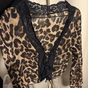 Leopardmönstrad blus - Snygg transparent blus med leopardmönster, svarta spetsdetaljer längs v-ringningen och snörning framtill. Långärmad och croppad modell som ger en edgy vibe. Perfekt för dig som vill sticka ut med djurmönster och coola detaljer.