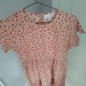 Blommig kortklänning från Name It - Söt klänning från Name It i ljus persikoton med små orange blommor över hela tyget. Klänningen har rund hals, korta ärmar och ett lätt rynkat midjeparti som ger en skön passform. Perfekt för sommarens alla tillfällen!