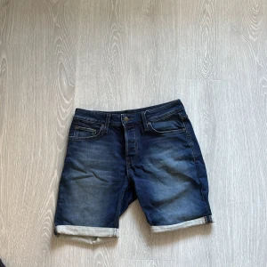 Blå jeansshorts från Jack & Jones - Snygga blå jeansshorts från Jack & Jones med klassisk femficksdesign och uppvikta benslut. Shortsen har normal passform, knapp och dragkedja framtill samt detaljerade sömmar på bakfickorna. Perfekta för en chill sommardag.