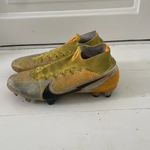 Nike fotbollsskor gul/svart - Säljer ett par Nike fotbollsskor med högt skaft i stickad syntet. Skorna är gula med inslag av grått och svart, har snörning och en stor svart Nike-logga på sidan. Sulan har fasta dobbar för gräsplan och detaljer i orange och svart. Perfekt för dig som vill sticka ut på planen. Men pris går att diskuteras eftersom ena skon är lite trasig på ens sida om ni ser på bilden