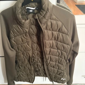Olivgrön pufferjacka från lager 157 - Säljer en olivgrön pufferjacka från 2117 med quiltad framsida och dragkedja. Jackan har långärmade släta ärmar och hög krage som ger extra värme. Perfekt för kyliga dagar och har en snygg, sportig look.