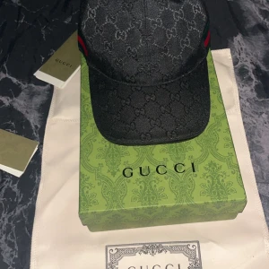 Svart keps från Gucci med GG-mönster - Snygg svart keps från Gucci med klassiskt GG-monogram över hela kepsen. Kepsen har grönt och rött band på sidorna och justerbar rem med baktill i skinn. Tillverkad i polyester och bomull, perfekt för dig som vill sticka ut med lyxig streetstyle. Pris går att diskutera helt ny och oanvänd 