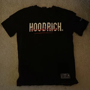 Svart Hoodrich t-shirt med tryck - Svart t-shirt från Hoodrich med stort vitt och rött tryck på bröstet och ryggen. Klassisk passform med korta ärmar och rund hals. Materialet är mjuk bomull och har coola detaljer som text på ärmen och nertill. Perfekt för streetwear-stilen. Mindre passform