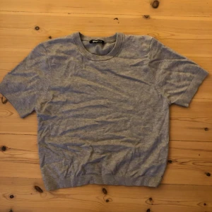 Grå stickad tröja från Gina Tricot - En basic grå stickad tröja från Gina Tricot i storlek S. Tröjan har rund halsringning, korta ärmar och ribbade muddar vid hals, ärmslut och nederkant. Perfekt för lager-på-lager och enkel att matcha med allt.