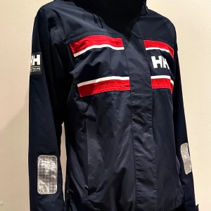Helly Hansen marinblå seglarjacka - Snygg marinblå vindjacka från Helly Hansen med röda och vita ränder på bröstet och ryggen. Jackan har huva, långa ärmar med reflexdetaljer och HH-logga på ärmen. Perfekt för dig som gillar sportig stil och vill sticka ut med coola färgblock.