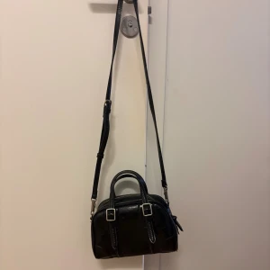 Svart axelväska från Zara - Snygg svart axelväska från Zara i skinnimitation med två silverfärgade spännen framtill. Väskan har både handtag och justerbar axelrem samt dragkedja upptill. Kommer inte till användning 🌸