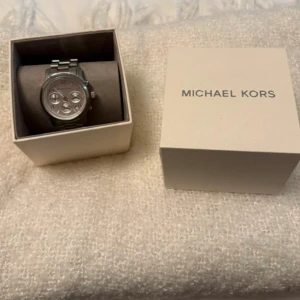 Silverfärgad klocka från Michael Kors - Säljer en stilren klocka från Michael Kors i silverfärgat metall med klassisk länkarmband. Klockan kommer i originalförpackning och har en rund urtavla med detaljerade knappar på sidan. Perfekt accessoar för dig som gillar snygga detaljer.