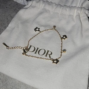 Guldarmband med Dior-charms - Supersnyggt guldarmband från Dior med tunna kedjor och små Dior-logga-charms i svart och guld. Armbandet har även glittriga stenar som detaljer och en justerbar kedja. Perfekt för dig som gillar lyxiga accessoarer med unik touch.