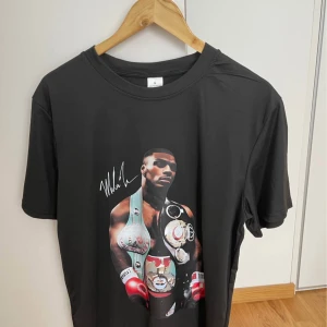 Svart t-shirt med boxare-tryck  - Svart t-shirt i 100% polyester med stort tryck av en boxare med bälten och röda handskar på framsidan. Snygg signaturdetalj vid motivet. Klassisk passform och rund halsringning, perfekt för dig som gillar sportiga och unika prints.