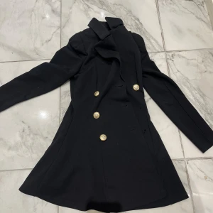 Svart rock med guldknappar - Snygg svart rock med markerad midja och långa ärmar. Dubbelknäppt design med sex guldiga knappar framtill som har ett coolt emblem. 2 styckna knappar har gått av , man kan dock sy in egna knappar. Jackan har klassisk krage och är tillverkad i ett slätt tyg som ger en stilren look.