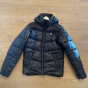 Mörkblå pufferjacka från G-Star RAW - Snygg mörkblå pufferjacka från G-Star RAW med huva och dragkedja framtill. Jackan har quiltad design, två fickor med knappstängning och en diskret logga på bröstet. Perfekt för kalla dagar när du vill hålla stilen. Fint skick, hör av dig vid frågor