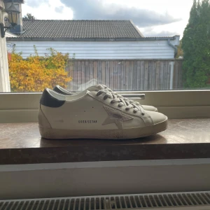 Golden Goose Super-Star sneakers vit/grå - Golden Goose Super-Star sneakers i vit skinn med grå detaljer och grå snörning. Ikonisk stjärna i mocka på sidan och grå häl med logotyp. Platt sula med vintage-inspirerad finish. Perfekt för dig som gillar en cool och avslappnad stil.