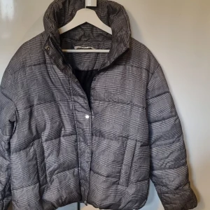 Rutig pufferjacka i svart och vitt - Säljer en snygg pufferjacka med rutigt mönster i svart och vitt. Jackan har hög krage, två stora fickor framtill och stängs med både dragkedja och tryckknappar. Tillverkad i 100% polyester, perfekt för kyliga dagar. Hällan har lossnat. Se sista bilden. Annars inget fel.