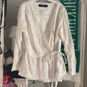 Vit omlottblus från Capri Collection - Supersöt vit omlottblus från Capri Collection med broderade blommor längs v-ringningen och knyt i midjan. Långärmad och lätt utsvängd nertill, perfekt för en fräsch och romantisk look. Broderad stjärna och logga på ryggen ger en extra detalj.