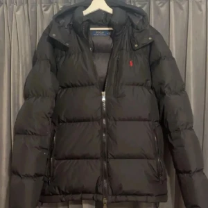  Ralph Lauren jacka  - Svart pufferjacka från Polo Ralph Lauren med huva och dragkedja framtill. Jackan har det klassiska röda Polo-logot på bröstet och är vadderad för extra värme. Perfekt för kalla dagar och har en clean, stilren look.