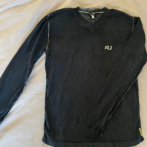 Svart stickad tröja från Armani Jeans - Svart stickad tröja från Armani Jeans med v-ringning och diskret AJ-logga broderad på bröstet. Långa ärmar och ribbade muddar ger en klassisk look. Perfekt för dig som gillar stilrena och enkla plagg med exklusiv känsla. Litet hål på axeln som går att sy igen ( ber man kan jag även göra det) nypris 1700kr 