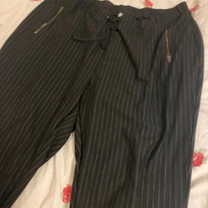 Svarta randiga byxor med snörning - Svarta byxor med tunna vita ränder, elastisk midja med snörning och dragkedjor vid fickorna. Sköna och stilrena med en avslappnad passform, perfekta för en trendig look. Materialet känns mjukt och lätt, och byxorna har en modern, lite loose fit.