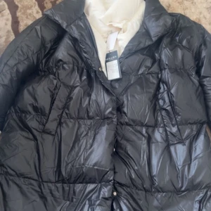 Svart lång dunjacka från Hollies - Lång svart dunjacka från Hollies med quiltad design och hög krage. Jackan har tryckknappar framtill och är fodrad i vitt. Perfekt för kalla dagar och har en stilren look som passar till det mesta.