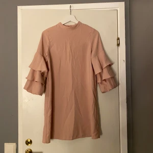 Beige kort klänning med volangärmar - Supersöt beige klänning med kort längd och högt halsringning. Ärmarna har två lager volanger som ger en extra cool och trendig look. Klänningen är rak i modellen och har en diskret dragkedja i ryggen. Perfekt för dig som gillar stilrena detaljer och voluminösa ärmar.💕ingen gratis frakt 