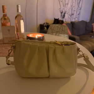 Beige/vit axelväska i skinnimitation - Snygg beige axelväska med rynkade detaljer och justerbar axelrem. Väskan har dragkedja upptill och guldfärgade metalldetaljer. Perfekt storlek för det viktigaste och passar till många olika outfits.