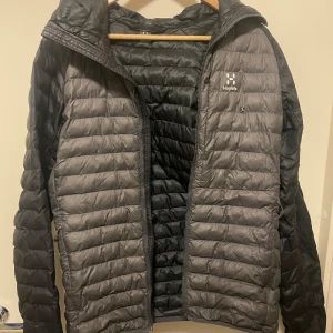 Svart quiltad dunjacka från Haglöfs - Snygg svart dunjacka från Haglöfs med quiltad design och huva. Jackan har dragkedja framtill och är lättviktig, perfekt för kyliga dagar. Den har en stilren look med diskret logga på bröstet och långärmade ärmar.