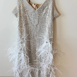 Glittrig kortklänning från Bella Barnett - Superglittrig kortklänning i champagnefärg från Bella Barnett, täckt av paljetter och pärlor. Klänningen har korta ärmar, v-ringning och är dekorerad med vita fjädrar och stora stenar nertill. Perfekt utklädsel för Halloween, tex 20-tal 