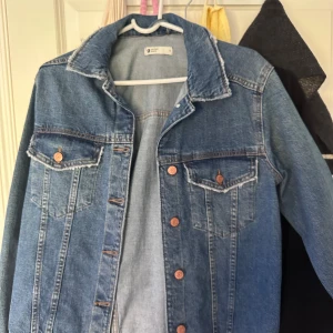 Klassisk blå jeansjacka från Gina Tricot - Säljer en klassisk blå jeansjacka från Gina Tricot med bronsfärgade knappar och två bröstfickor med lock. Jackan har en rak passform, krage och tydliga sömmar som ger en tidlös look. Perfekt för lager-på-lager och enkel att styla.