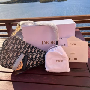 Dior Saddle Bag i canvas och skinn - Säljer en ikonisk Dior Saddle Bag med monogrammönster i mörkblått och beige canvas, detaljer i svart skinn och guldiga metalldetaljer. Väskan har justerbar axelrem och stort D-hänge framtill. Perfekt statement piece för dig som vill sticka ut.