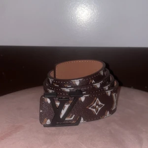 Brunt bälte med monogram - Snyggt brunt bälte med klassiskt monogrammönster i vitt och beige. Bältet har en svart logga som spänne och är gjort i präglat skinn. Perfekt accessoar för att lyfta din outfit.