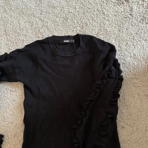 Svart stickad tröja med volang från Bik Bok - Svart stickad tröja från Bik Bok med rund halsringning och snygga volangdetaljer längs ena ärmen. Perfekt för dig som vill ha en basic tröja med en twist. Mjuk och skön att bära, passar till allt.
