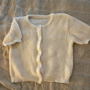 Vit stickad kofta med kort ärm - Superfin vit stickad kofta med korta ärmar och rund hals. Koftan har ribbad struktur och knäpps med små vita knappar framtill. Perfekt att slänga över en topp för en chill look. Passar dig som gillar mjuka och neutrala färger.