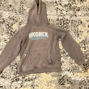 Grå hoodie från Hoodrich med tryck - Säljer en grå hoodie från Hoodrich med stor huva och tryck både fram och bak. Tröjan har en magficka med dragkedja och detaljer i ljusblått. Mjuk insida och klassisk streetstil, perfekt för dig som gillar snygga loggor och chill vibe.